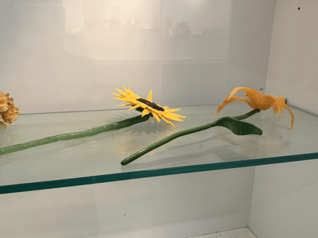 <b>Flower Models</b><br>The model originates from the Sheridan Crafts and Design building.This arrangement features models of flowers made from smooth, shiny materials that imitate natural petals. The bright yellow sunflower and delicate orange orchid contrast beautifully against the green stems, creating a vibrant and lifelike appearance. The glossy finish enhances the visual interest, mimicking the way sunlight would reflect off real flowers.