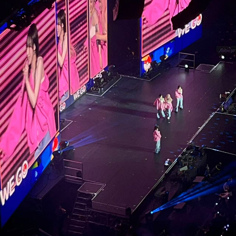 An indoor concert scene with performers on stage and large screens displaying the action, capturing the loud music and cheering crowd at a live performance.