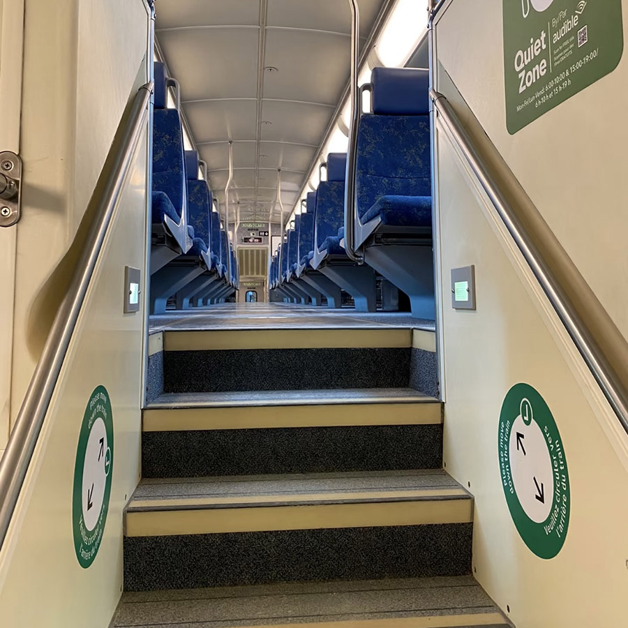 pThe interior of a train showing a staircase with 'Quiet Zone' signs, evoking the clear, mechanical voice announcing stops over the train’s speaker system.
