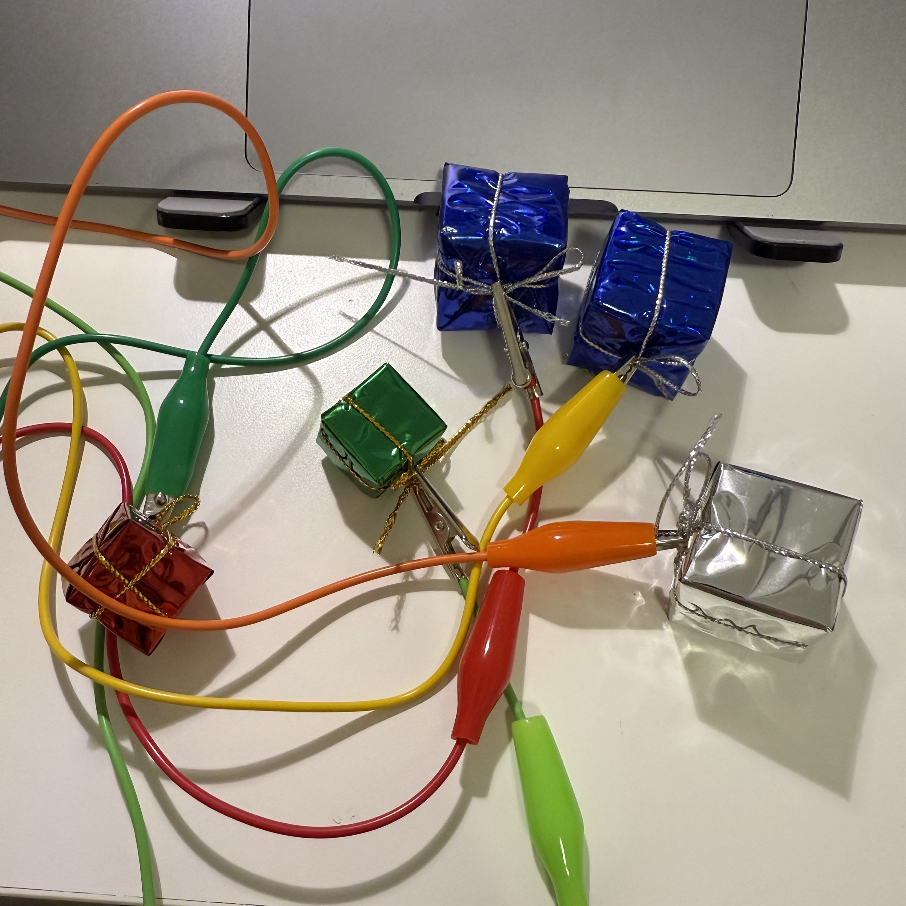 I have connected colorful gift-wrapped boxes to my Makey Makey board using alligator clips. Each box is wrapped in shiny paper and acts as a touch-sensitive button. When I touch these boxes, they send signals to the computer, allowing me to interact with digital applications in a fun and festive way.