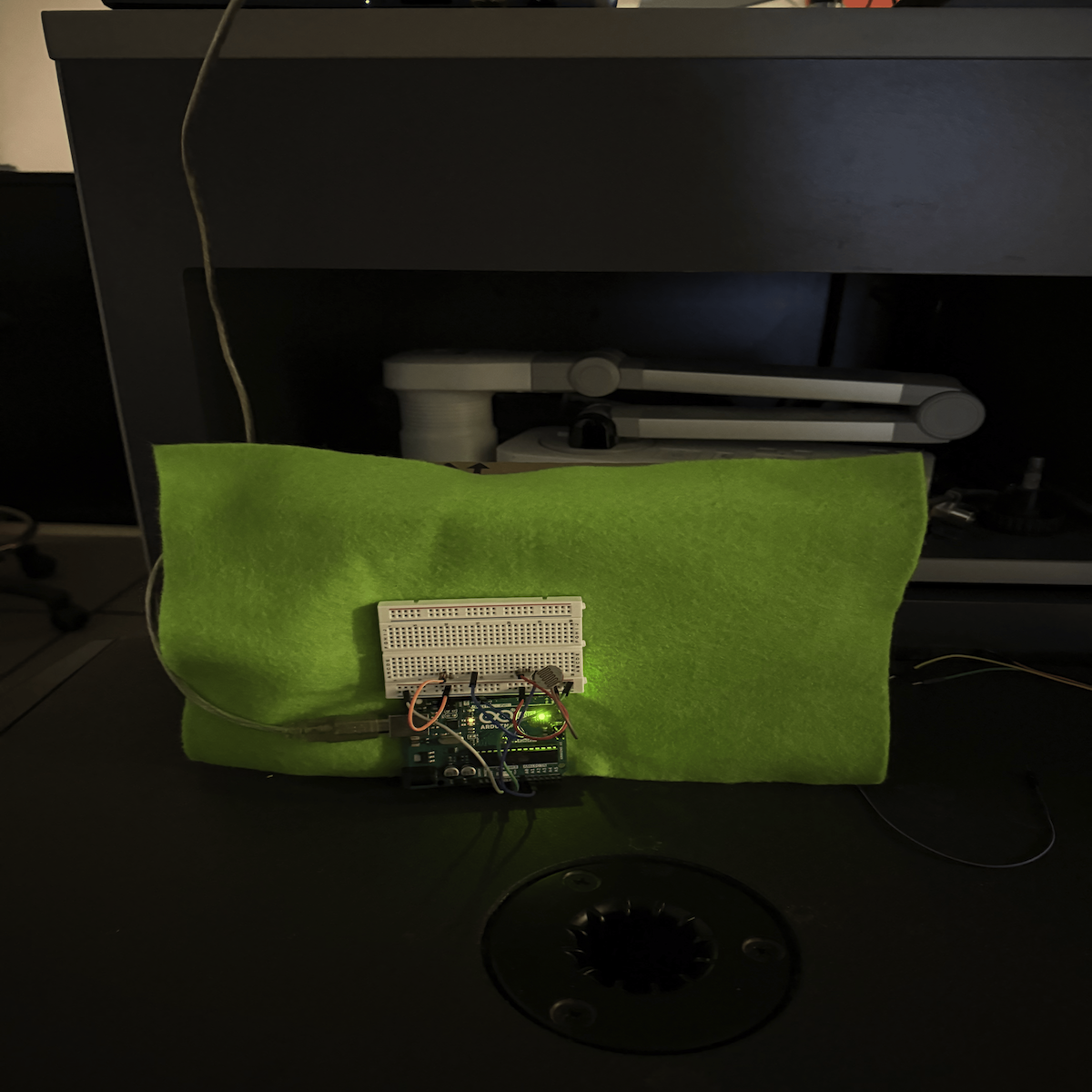 <b>Testing the Circuit Behind the Fabric</b><br>We placed the Arduino and breadboard in font of a piece of fabric to simulate the cushion surface. The setup allowed us to check how the sensor reacted when the light changed in front of the fabric. This was our first step in building the physical prototype.